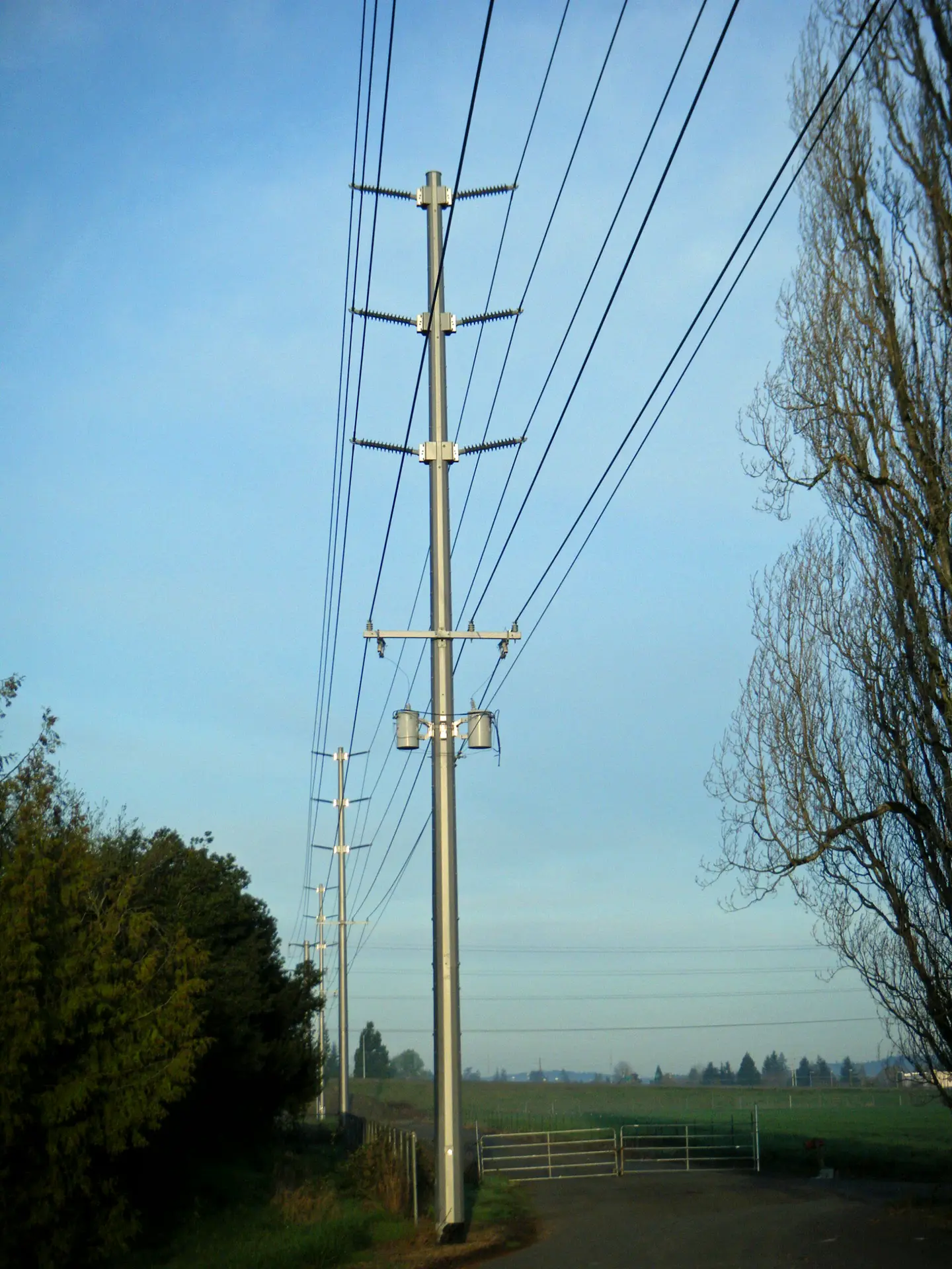 Galvanized steel distribution pole with crossarms and transformers beside a field